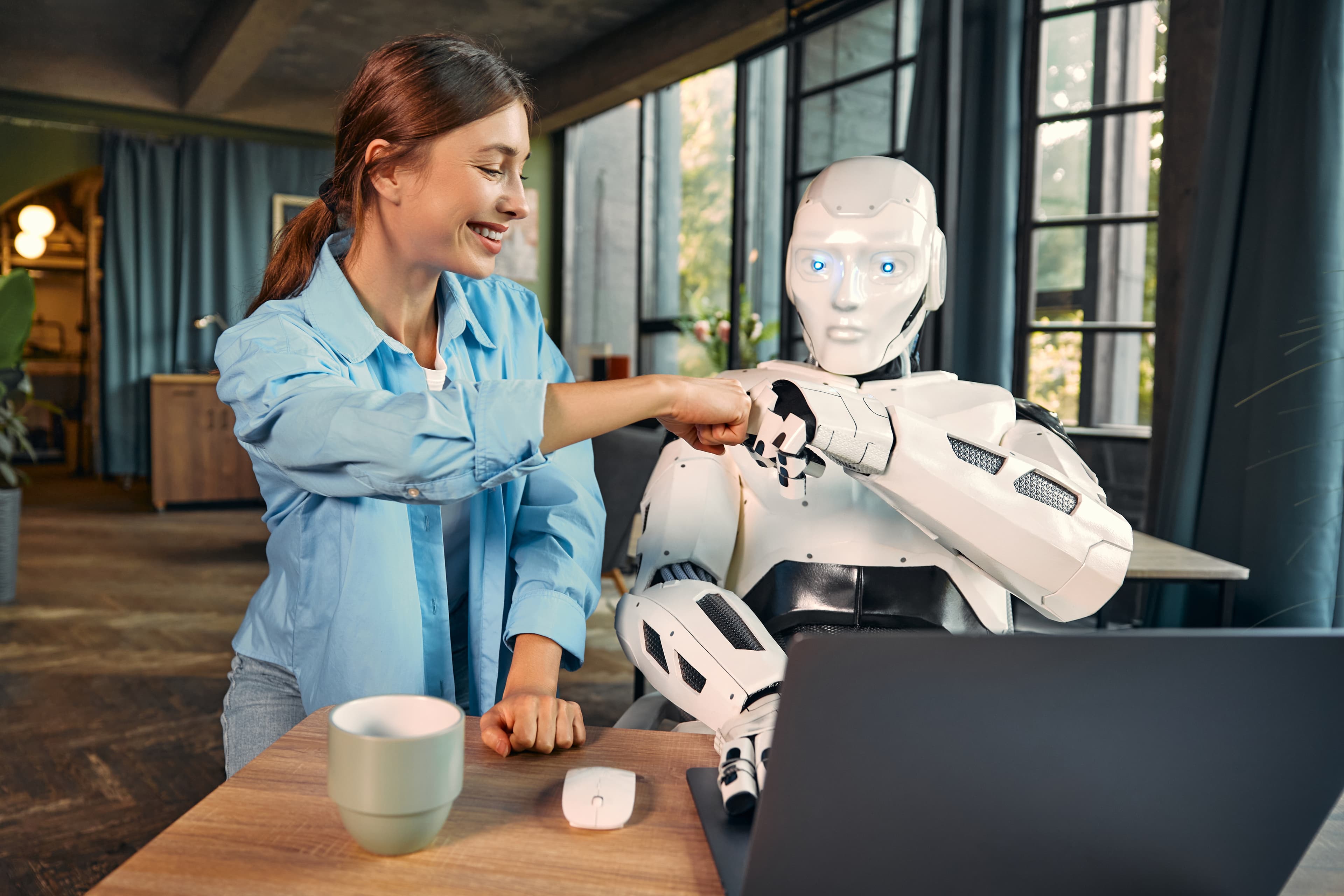A woman and a humanoid robot bumping fists across a desk